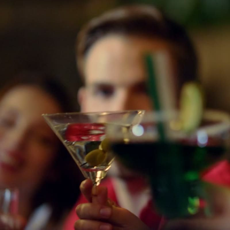 A martini glass with olives on a pick held up, the room soft behind it